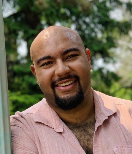 Travis Leon, tenor, smiling in a light pink shirt in an outdoor setting.