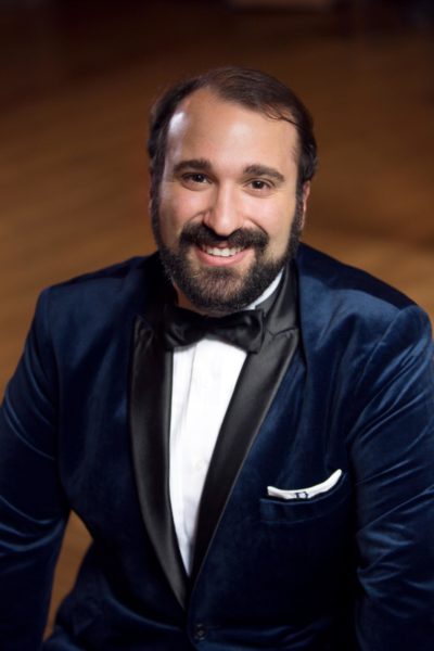 Christopher Bozeka, a friendly face with facial hair, he is smiling in a blue velvet tuxedo.