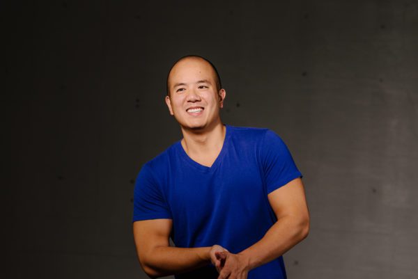 André Chiang, baritone, smiling with hands clasped against a dark background, he is wearing a vibrant blue shirt.