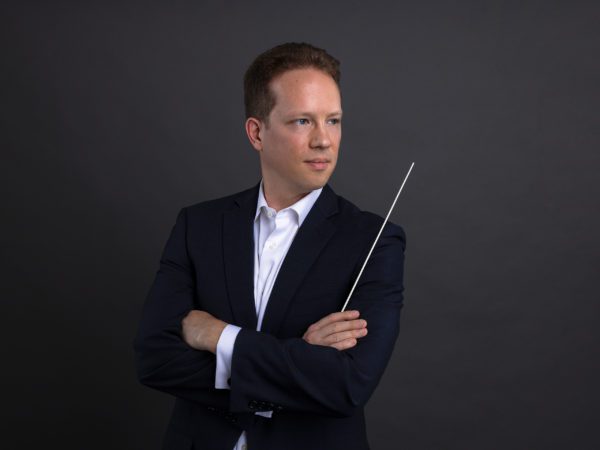Joshua Horsch, posting with arms crossed in a dark suit and his conducting wand in one hand.