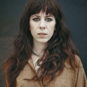 Missy Mazzoli, looking straight at the camera with dramtic makeup and expression, auburn hair and a brown muslin top.