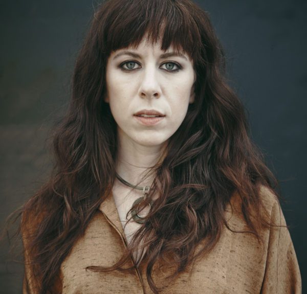 Missy Mazzoli, looking straight at the camera with dramtic makeup and expression, auburn hair and a brown muslin top.