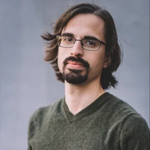 Evan L. Snyder, standing casually in a dark green cashmere sweater and jeans. He has long dark hair, glasses, and facial hair.
