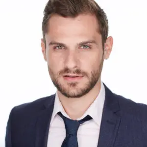 Brian James Myer, a handsome young man with short facial hair in a loosely tied tie and suit, against a white background.
