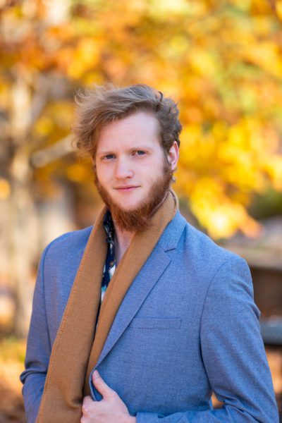 Joshua Hager, an auburn-haired gentleman with a long beard, wearing a light blue suit and a brown scarf in an autumn setting.