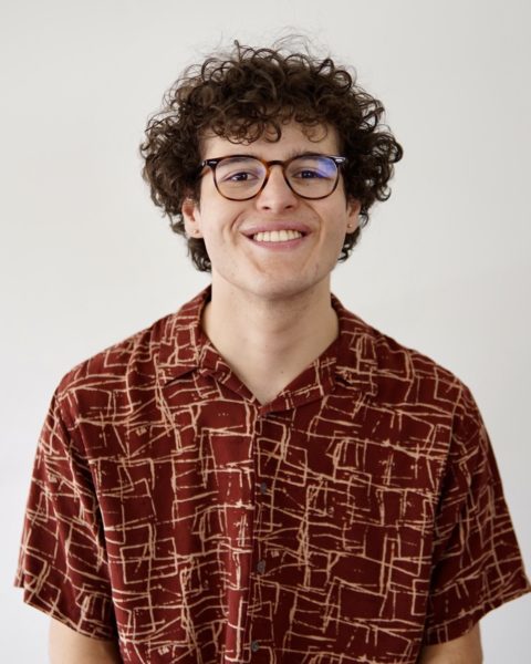 Rafael Canizalez, with glasses and shaggy curly hair in a bold-patterned shirt, smiling against a plain background.
