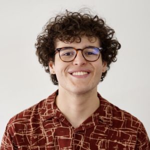 Rafael Canizalez, with glasses and shaggy curly hair in a bold-patterned shirt, smiling against a plain background.