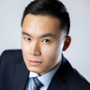 Operatic Tenor Michael TK Lam, a young Asian man with a neutral expression wearing a suit and tie, tightly-cropped headshot against a light grey background.