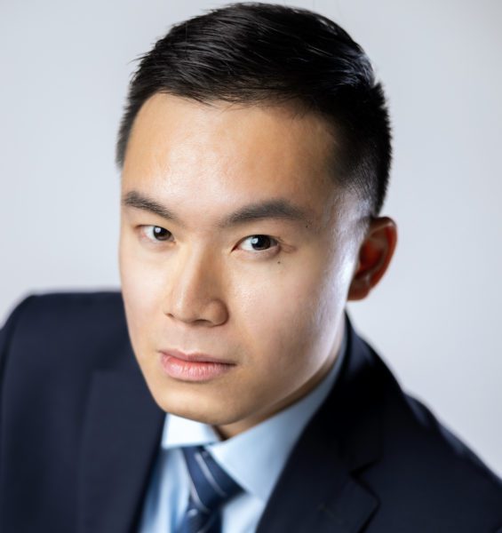 Operatic Tenor Michael TK Lam, a young Asian man with a neutral expression wearing a suit and tie, tightly-cropped headshot against a light grey background.