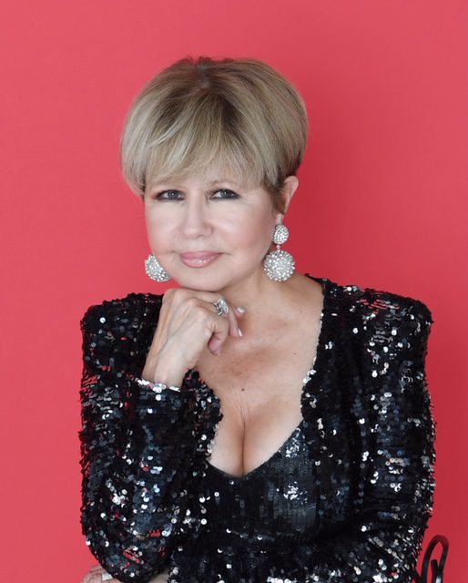 Pia Zadora wears a black sequined top and casually rests her chin on her fist as she stands against a bright red background.