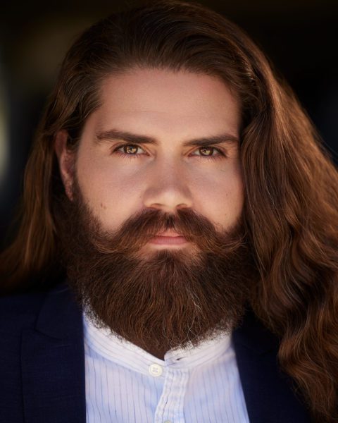 Arnold Geis, a young man with long hair and a full beard, close-up in a black suit.