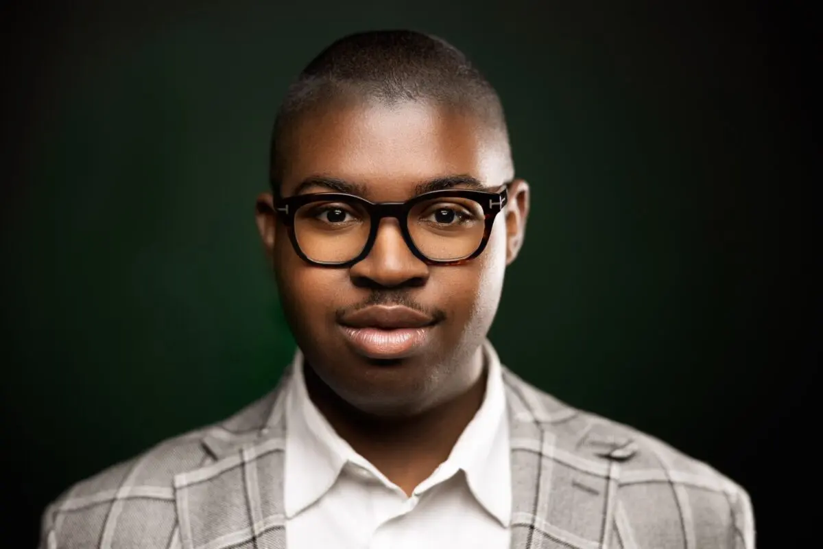 Christian Shelton, a young black man with dark-rimmed glasses and a tan plaid suit stands against a black background.