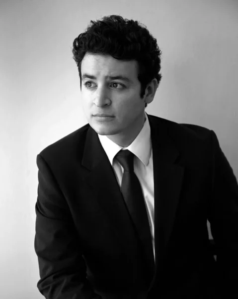 Daniel Montenegro, tenor, black and white photo. He wears a simple black suit with a white shirt and black tie.