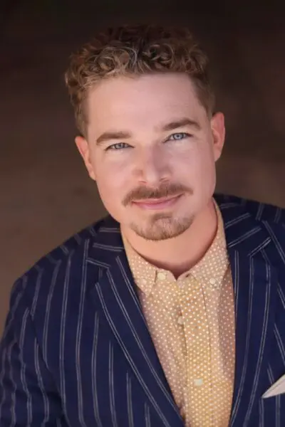 Joshua Hughes, headshot in a tan shirt and pin-striped suit.