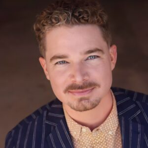 Joshua Hughes, headshot in a tan shirt and pin-striped suit.