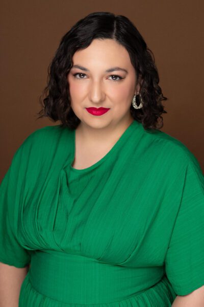 Claudia Affan, a light-skinned woman with shoulder-length dark hair and formal makeup in a professional studio photo. She wears a bright green loose-fitting blouse.