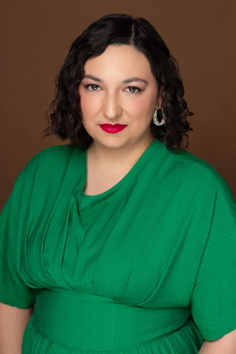 Claudia Affan, a light-skinned heavyset woman with shoulder-length dark hair and formal makeup in a professional studio photo. She wears a bright green loose-fitting blouse.