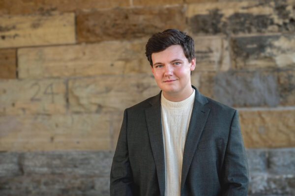 Justin Burgess, a young white dark-haired man with a clean-shaven face stands in a casual day suit in front of a slate brick wall.