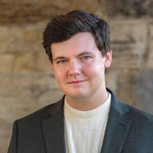 Justin Burgess, a young white dark-haired man with a clean-shaven face stands in a casual day suit in front of a slate brick wall.