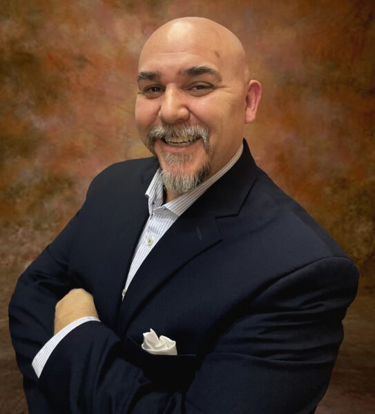 Adelmo Guidarelli, a bald man with a greying goatee, smiles and poses with arms crossed in a formal suit.