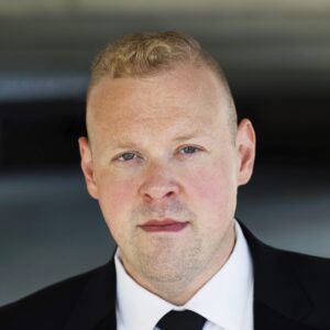 Ben Gulley with a neutral expression in a formal black suit and tie.