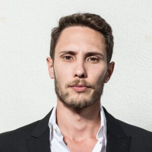 Colin Ramsey, a Chinese-American man with light facial hair, poses in a dark suit and a neutral serious expression.