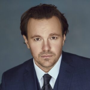 Daniel Scofield dressed in a suit and tie with a neutral expression and studio lighting.