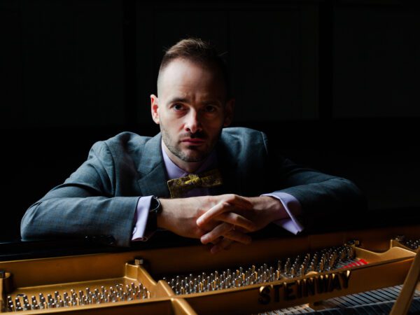 Nathan Carterette poses with a serious expression in dim, dramatic lighting as he rests on the soundboard of a grand piano.