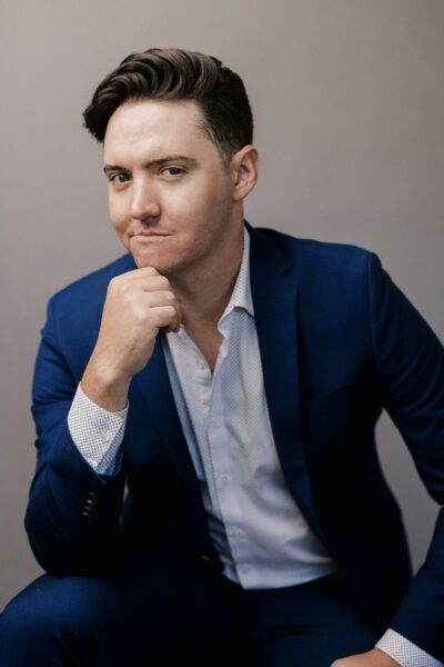Matthew Peterson, a young caucasian man, poses seductively in a formal suit with one hand near his chin.
