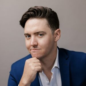 Matthew Peterson, a young caucasian man, poses seductively in a formal suit with one hand near his chin.