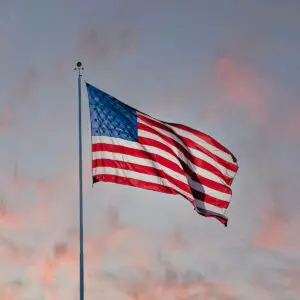 Opera Las Vegas' poster for "Voices Raised for Vets," featuring a solo pianist playing a grand piano in the middle of a field of wheat, symbolizing amber waves of grain. A flagpole with an American flag raised stands behind the piano, and the setting sun offers a calm, peaceful mood.