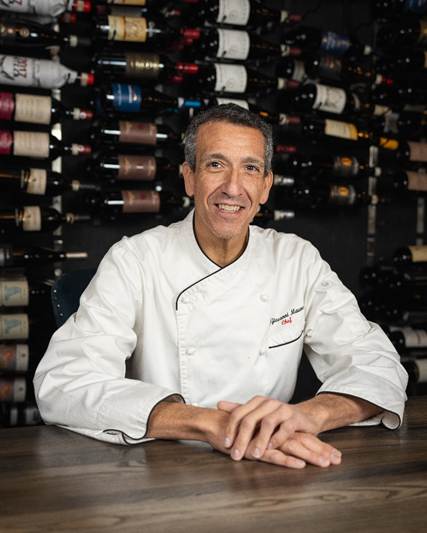 Chef Giovanni Mauro sits comfortably in his restaurant attire in front of a rack of wine bottles.
