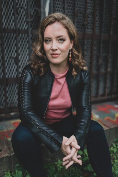 Katherine Whyte sits on a ledge in casual attire, under professional lighting and with a friendly but neutral expression.