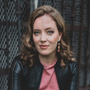 Katherine Whyte sits on a ledge in casual attire, under professional lighting and with a friendly but neutral expression.
