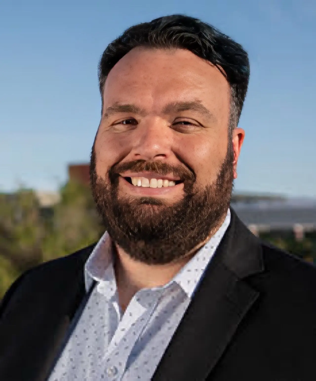 Jonathan Baltera, a tall, bearded man, smiles outdoors in sunlight. He wears a dark suit jacket and a light collared shirt without a tie.
