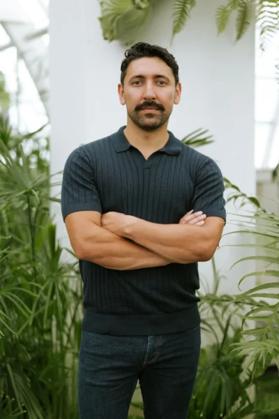 Efrain Solis stands with arms crossed, outdoors amid verdant greenery in casual attire.
