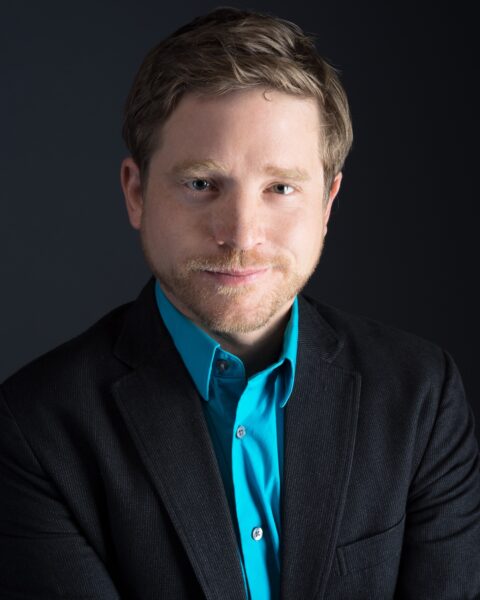 Jon Lee Keenan, a handsome man with short-cropped facial fair smiles gently in studio lighting. He wears a festive collared shirt and a dark formal suit.