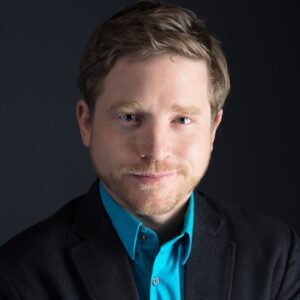 Jon Lee Keenan, a handsome man with short-cropped facial fair smiles gently in studio lighting. He wears a festive collared shirt and a dark formal suit.