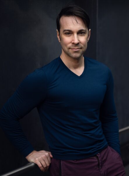 John de los Santos stands in dark, dramatic lighting wearing casual attire.