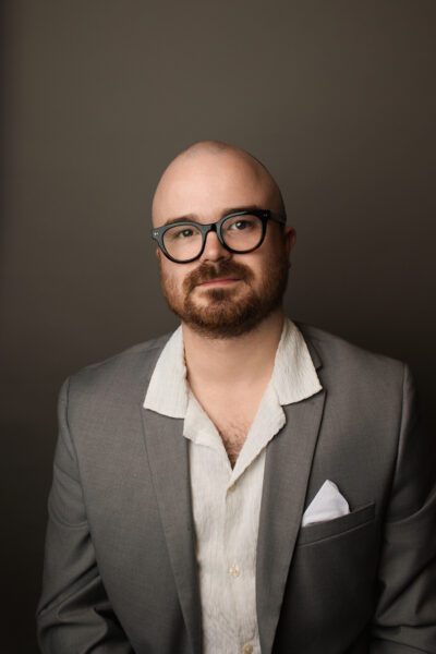 Spicer Carr headshot, a bald, bearded man with glasses in studio lighting and a casual sportcoat.