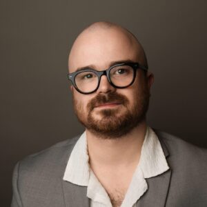 Spicer Carr headshot, a bald, bearded man with glasses in studio lighting and a casual sportcoat.