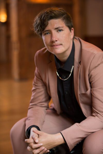 Rose Freeman sits casually resting her elbows on her knees. She wears a light brown day suit over a black tee.